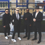 Tonic Sol-Fa, a cappella quartet group, stands in black suits on a building overlooking other buildings.